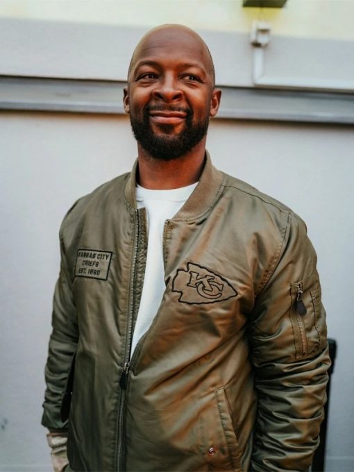 Man wearing a green bomber jacket with a logo, standing against a light-colored wall in UK